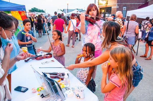 A violinist plays on the street in Benson while children do a craft activity at a table.